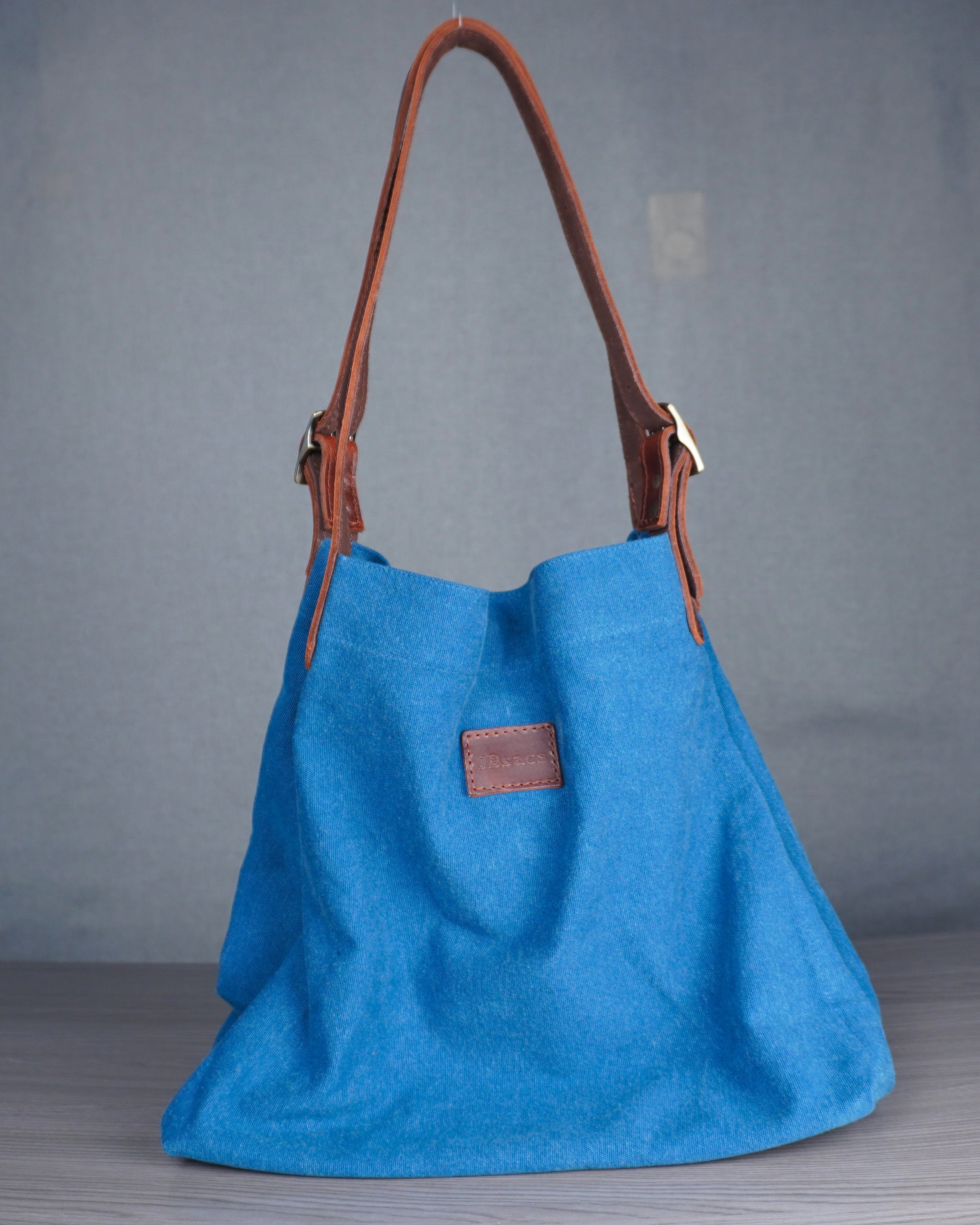 Blue handbag with brown leather handle on a gray floor against a blue wall.