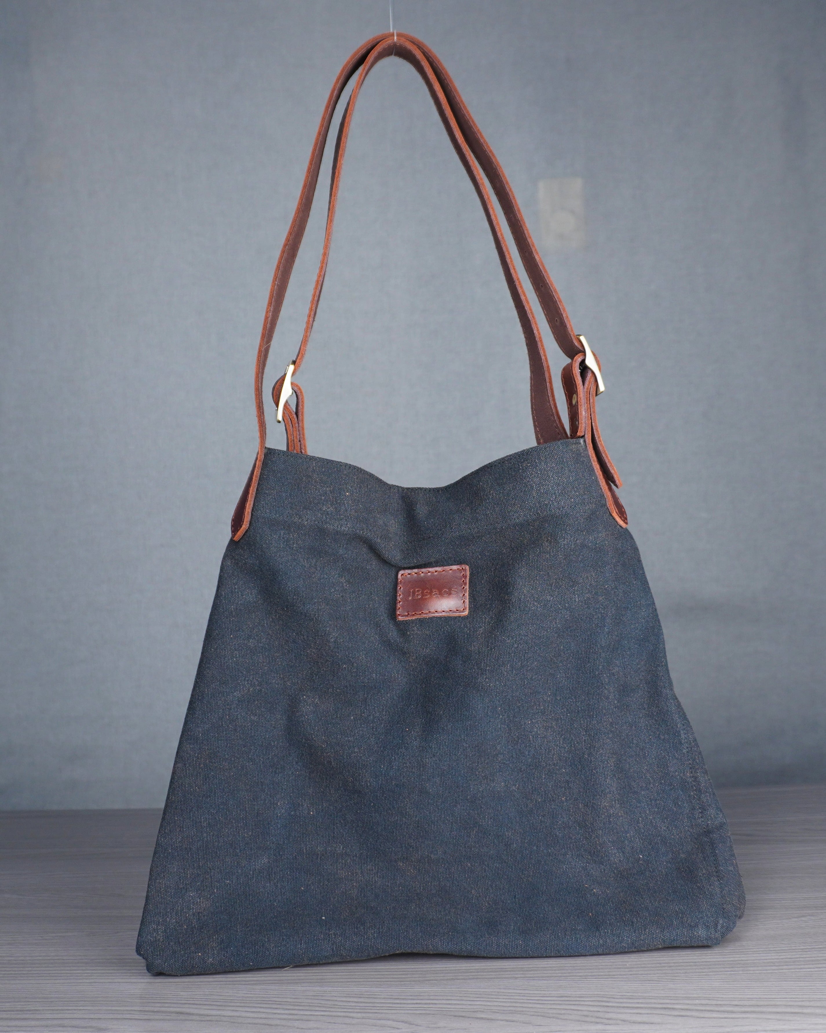 Gray handbag with brown leather handle and strap on a wooden floor against a gray wall.