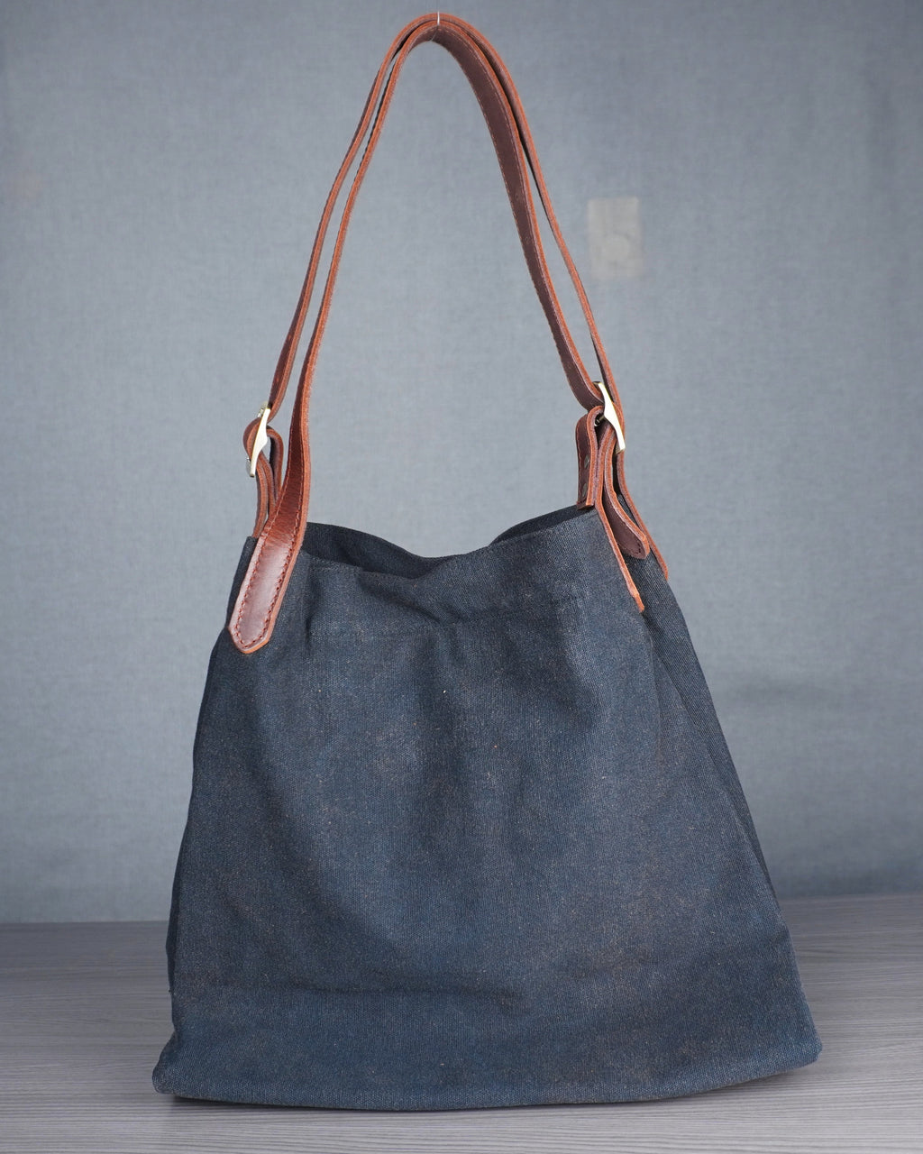 Dark gray handbag with brown leather handle on a wooden floor against a blue wall.