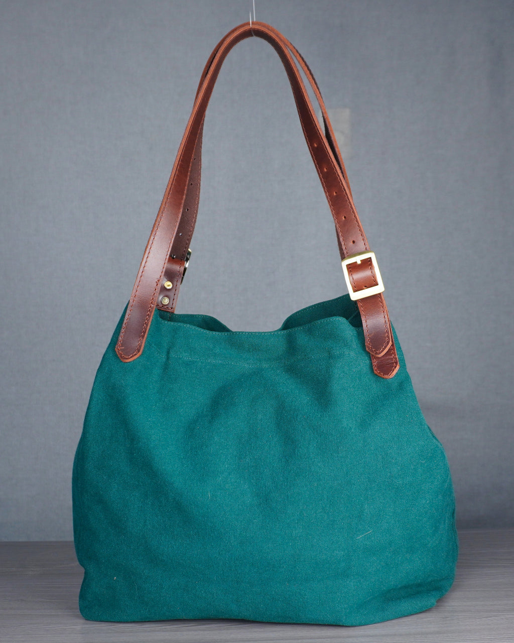 Green handbag with brown leather handles on a wooden floor against a gray wall.