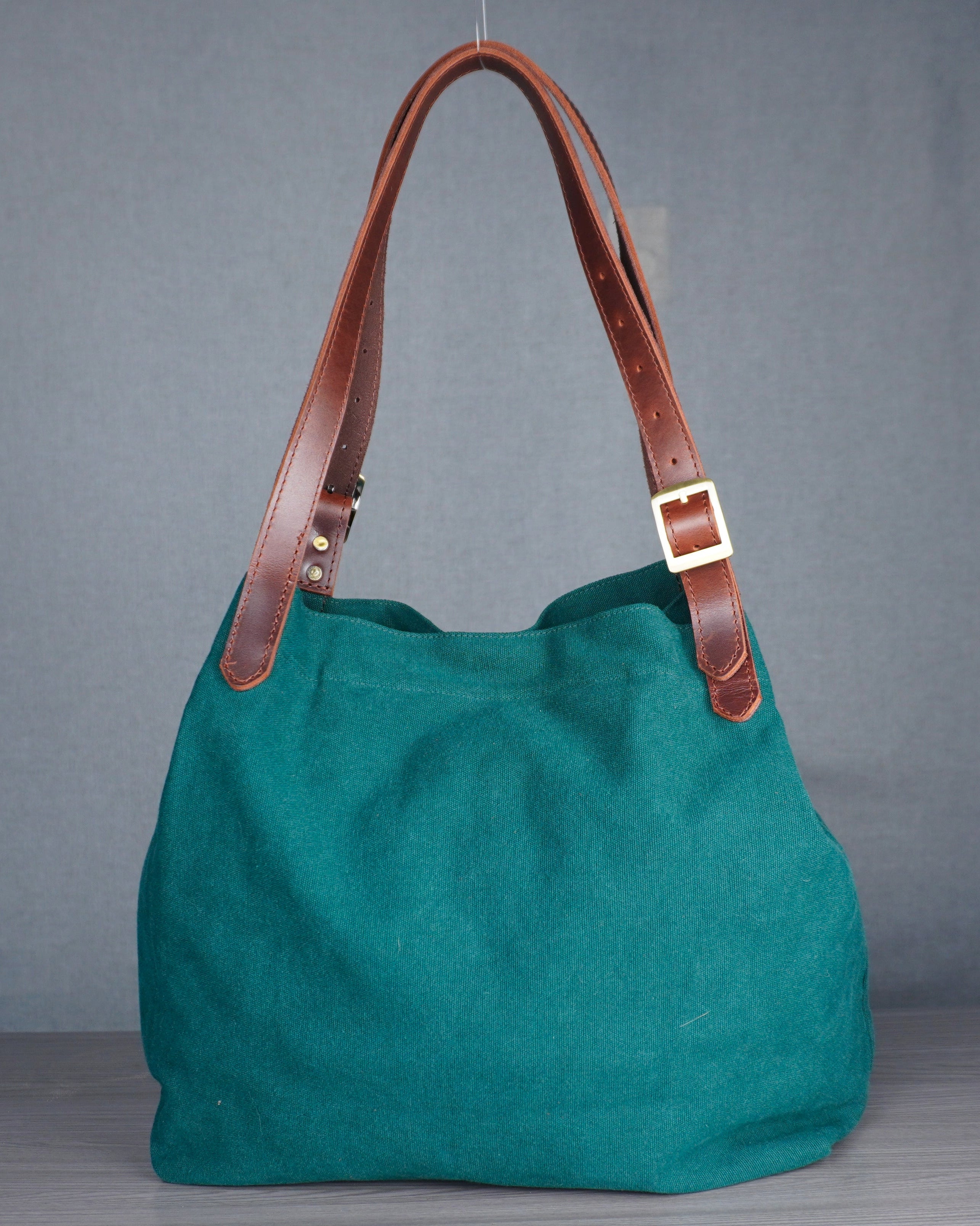Green handbag with brown leather handles on a wooden floor against a gray wall.