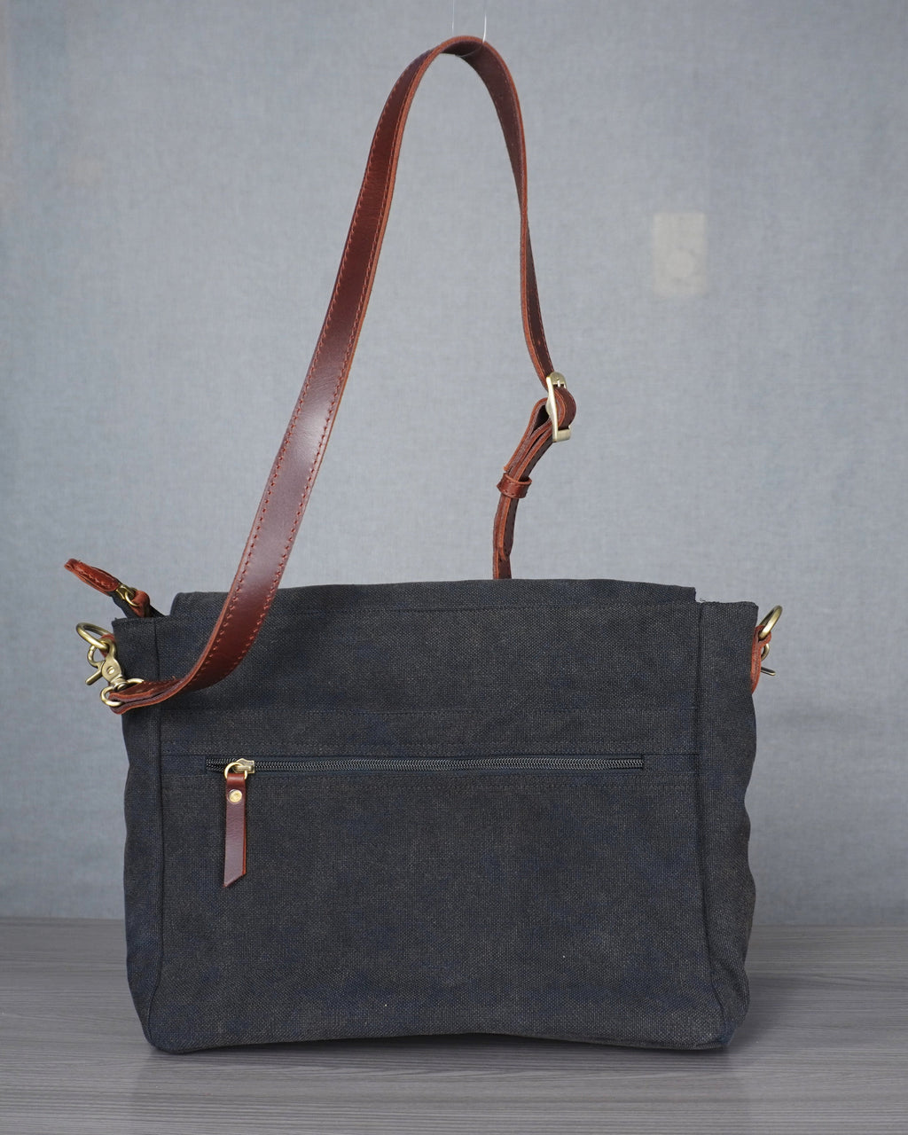 Gray handbag with a brown leather strap on a gray surface against a blue and gray wall.